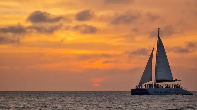 A sailing catamaran of passengers silhouetted against an orange sunset over the sea